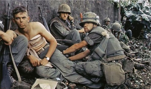 Foto van een Amerikaanse soldaten die zich schuilhouden achter een muurtje. De gewonde word verzorgt door de dokter 
